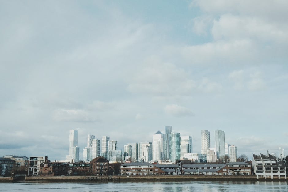 A wide view of a modern city skyline featuring numerous high-rise office buildings and skyscrapers with glass facades reflecting the daylight, situated behind a shoreline with lower residential and co
