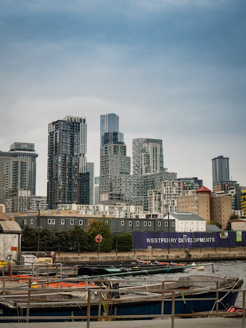 A wide view of a modern city skyline featuring numerous high-rise office buildings and skyscrapers with glass facades reflecting the daylight, situated behind a shoreline with lower residential and co