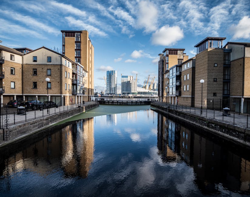 A narrow canal or waterway is flanked by modern, multi-storey residential buildings constructed with light brown brickwork and dark metal-framed windows. The buildings have clean, contemporary lines, 