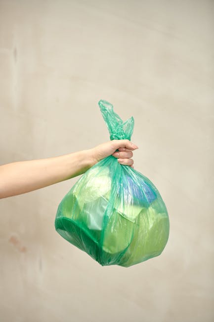 A large green waste disposal bin positioned outdoors on a paved surface, placed against a dark brick wall. The bin is filled with black and red plastic bags and some packaging, likely containing garde