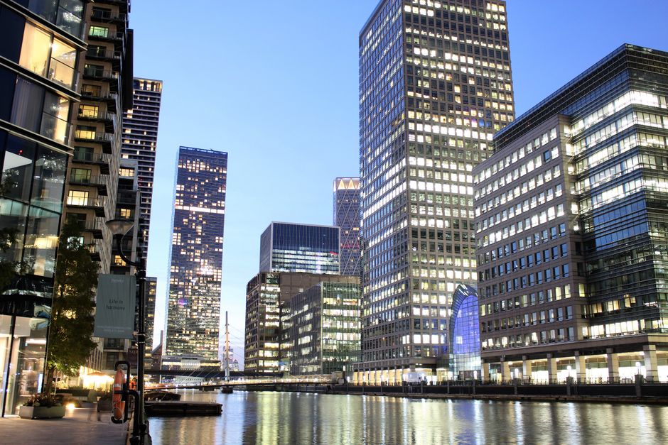 The image depicts a modern urban scene viewed from beneath a dark, concrete archway or bridge structure, leading out onto a paved area. In the foreground, the underside of the structure is composed of